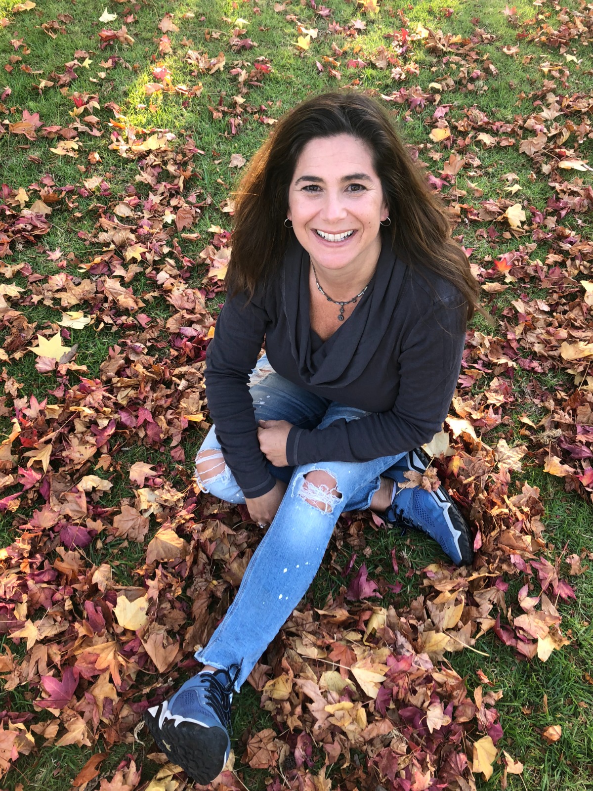 Cara J Stevens author wearing black shirt and distressed jeans sitting in a pile of leaves outdoors.